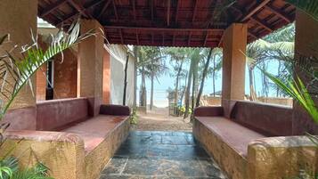 A covered outdoor seating area with stone flooring and wooden benches, surrounded by palm trees and a view of the beach.