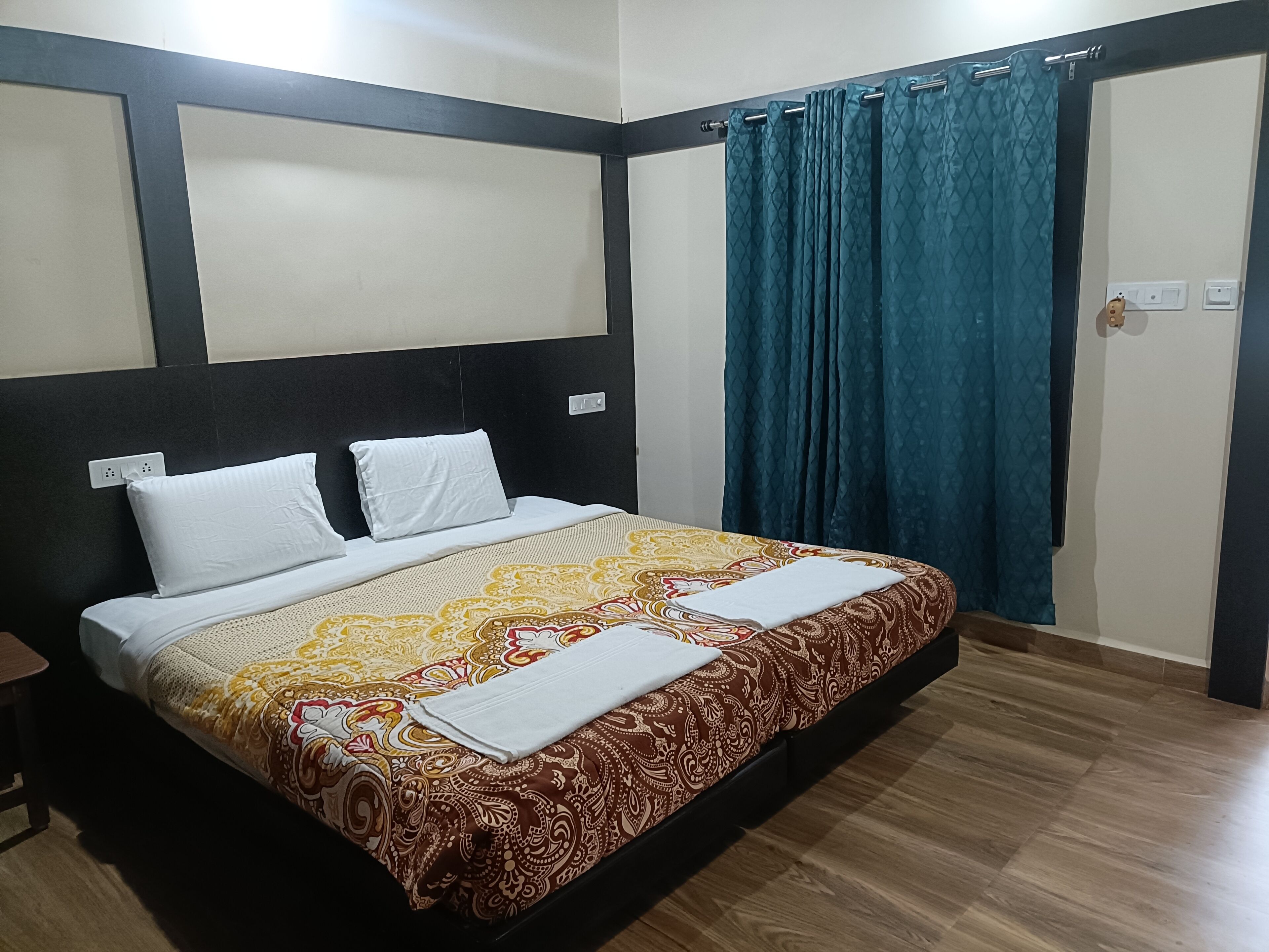 A hotel room with a large bed, two white pillows, a patterned bedspread, a blue curtain, and a wooden floor.