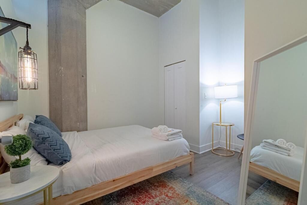 Ein Schlafzimmer mit einem Bett, einem kleinen Tisch mit Pflanze, einem Spiegel, einem Kleiderschrank und einer Lampe.