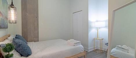 Ein Schlafzimmer mit einem Bett, einem kleinen Tisch mit Pflanze, einem Spiegel, einem Kleiderschrank und einer Lampe.