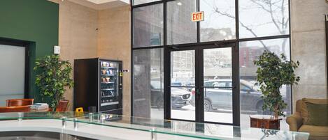 A lobby with a glass partition, vending machine, potted plants, and a view of the street outside.