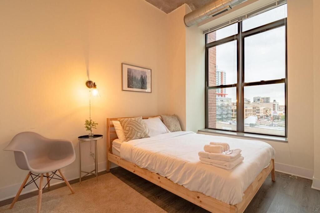A bedroom with a bed, a chair, a bedside table, a window with a city view, and a picture on the wall.