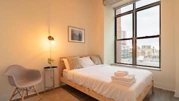 A bedroom with a bed, a chair, a bedside table, a window with a city view, and a picture on the wall.