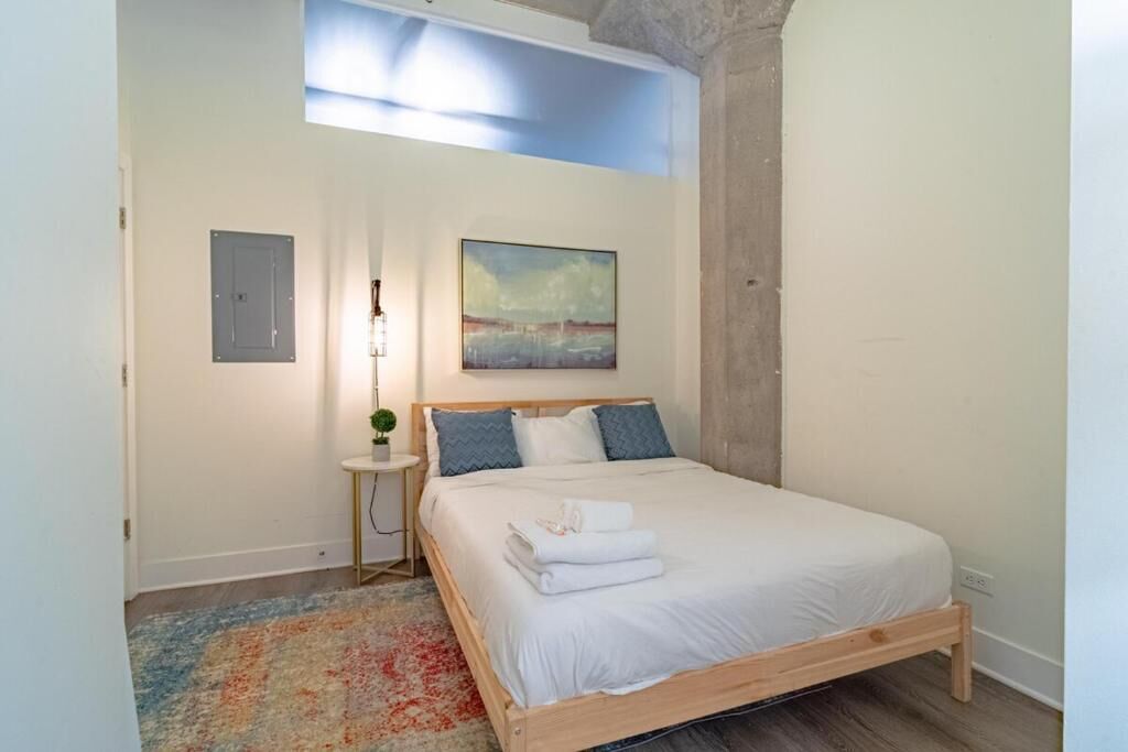 Una camera da letto con un letto, un comodino, un tavolino di fianco al letto, un quadro appeso e una finestra.