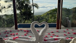 A bed with a heart-shaped towel arrangement and rose petals, set against a backdrop of a scenic view through large windows. - DRAKO VILLAS (San Juan)