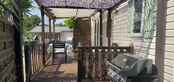 Een overdekt terras buiten met een barbecue, stoelen en een tafel.