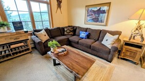 Un salon chaleureux avec un canapé d’angle, une table basse en bois et une grande fenêtre donnant sur l’extérieur.