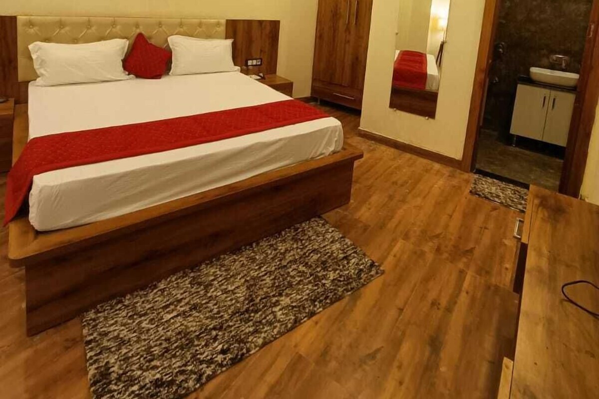 Quarto com cama grande, cabeceira de madeira, manta vermelha, mesa de cabeceira de madeira, guarda-roupa, ar condicionado e um banheiro pequeno com pia e espelho.