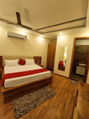 A bedroom with a large bed, wooden headboard, a red throw blanket, a wooden nightstand, a wardrobe, an air conditioning unit, and a small bathroom with a sink and mirror. - Hotel Moon Regency (Panipat)