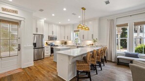 A modern kitchen with white cabinetry, stainless steel appliances, a central island with bar stools, and a welcoming sign.
