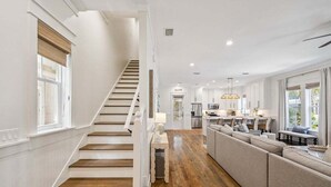A spacious living area with a wooden staircase, a kitchen in the background, and a large window providing natural light.