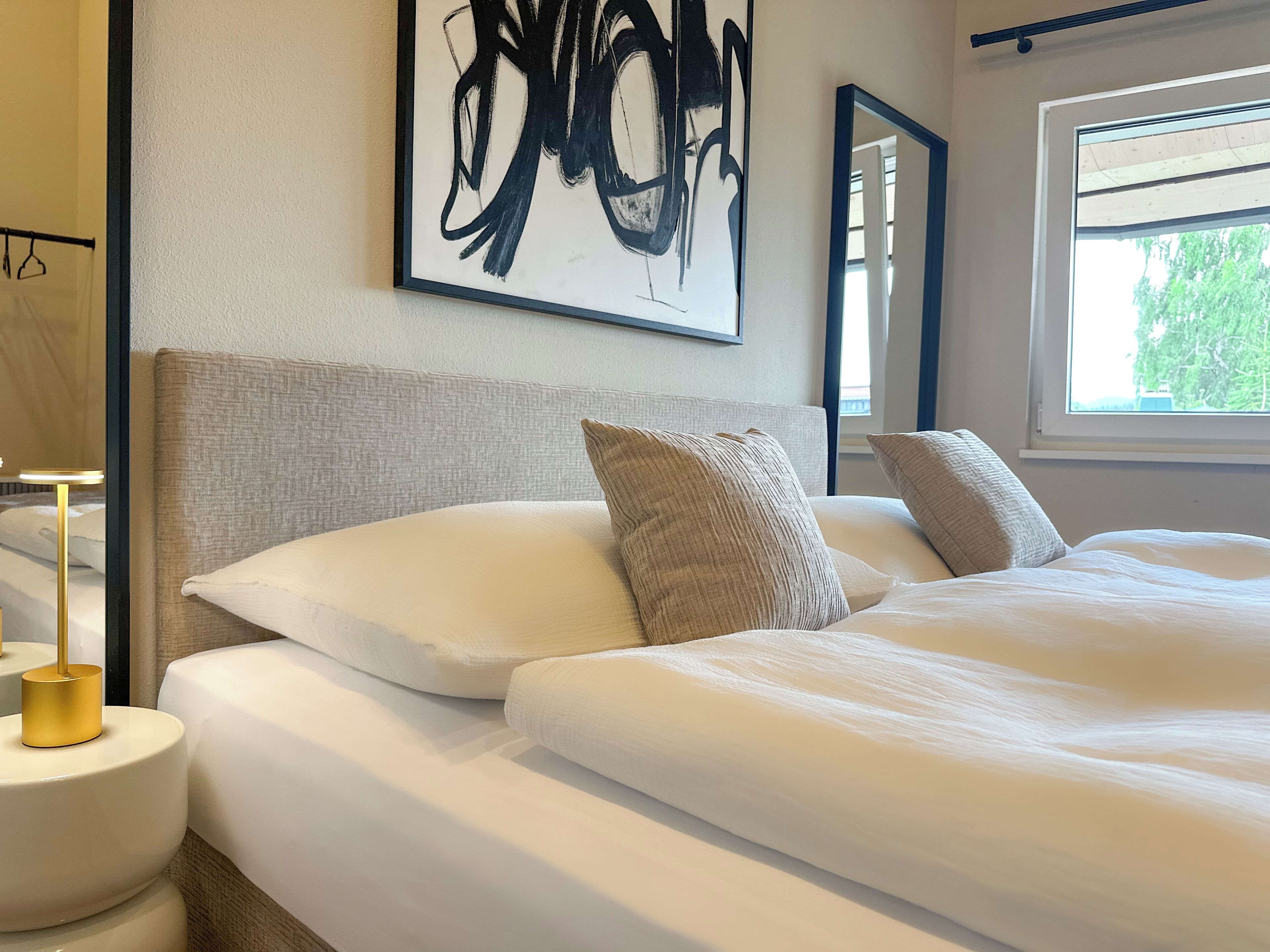 Een netjes opgemaakt bed met witte lakens en twee kussens, een nachtkastje met een lamp, en een groot abstract schilderij aan de muur.