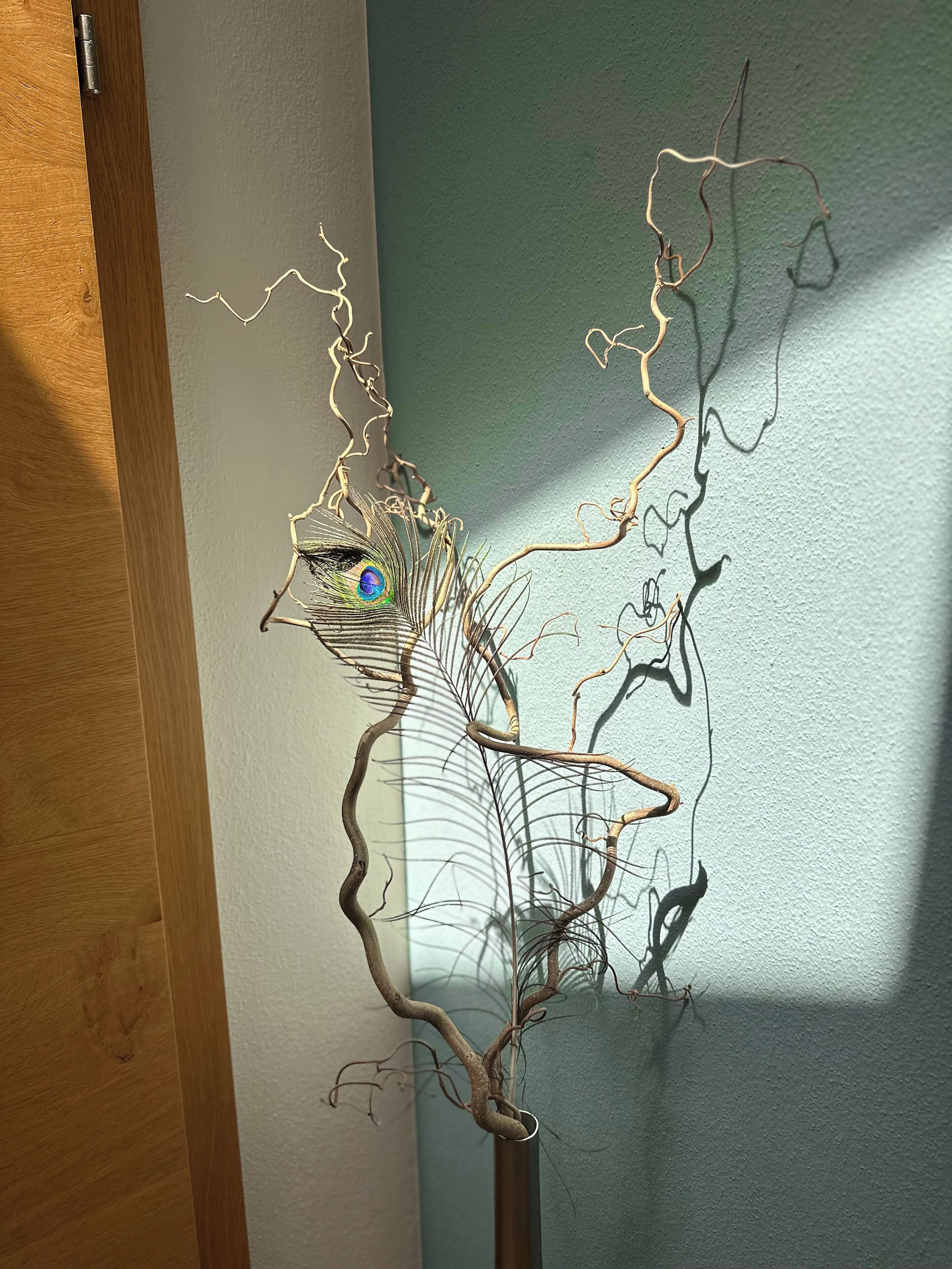 Metallinen veistos, joka esittää puuta, ja jonka oksalla on lintu. Veistos on sijoitettu seinää vasten.