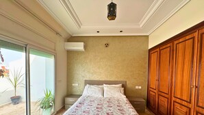 A bedroom with a large bed, a wardrobe, a ceiling light, and a sliding glass door leading to an outdoor area with plants.