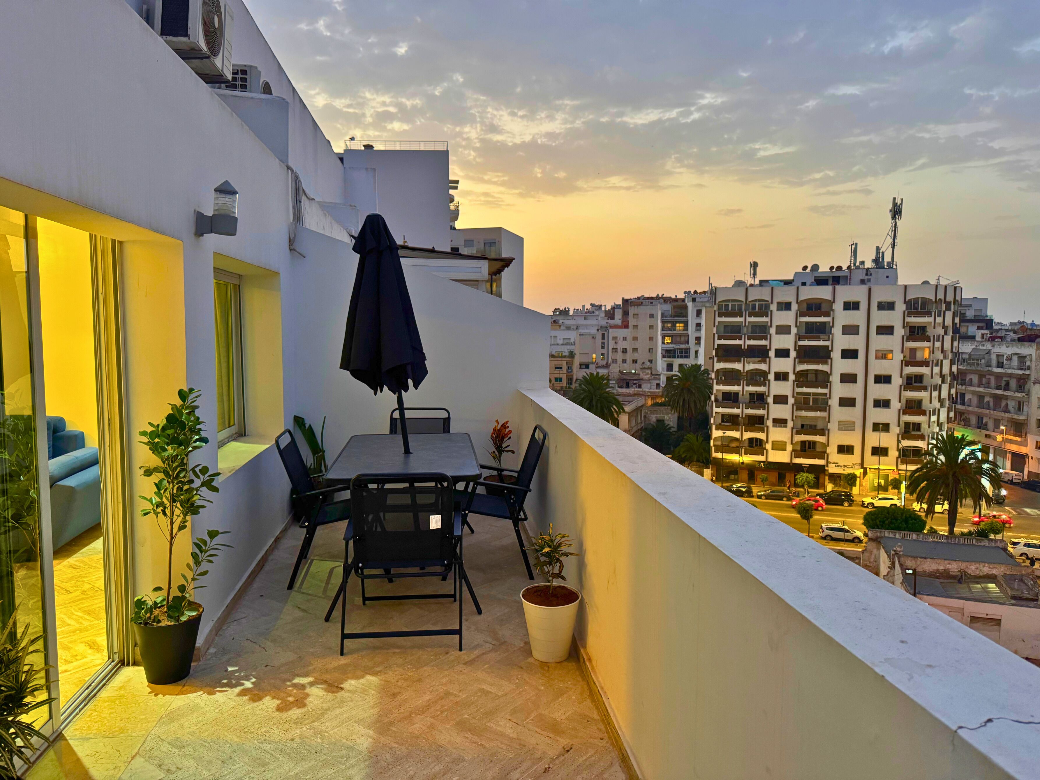 Un balcón con mesa y sillas, plantas en macetas y vistas a la ciudad al atardecer.