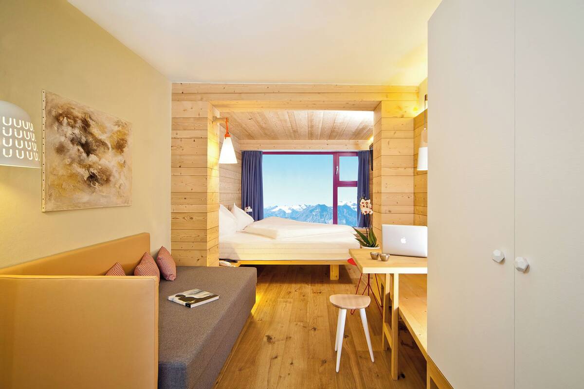 A hotel room with a bed, a desk with a laptop, a small stool, a window with a mountain view, and a painting on the wall.