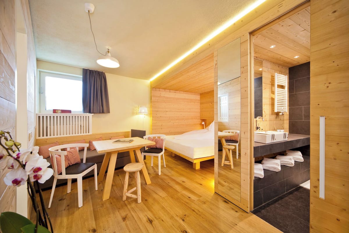 A cozy room with a wooden floor, a bed, a desk, a chair, a small stool, and a bathroom with a sink and towel rack.