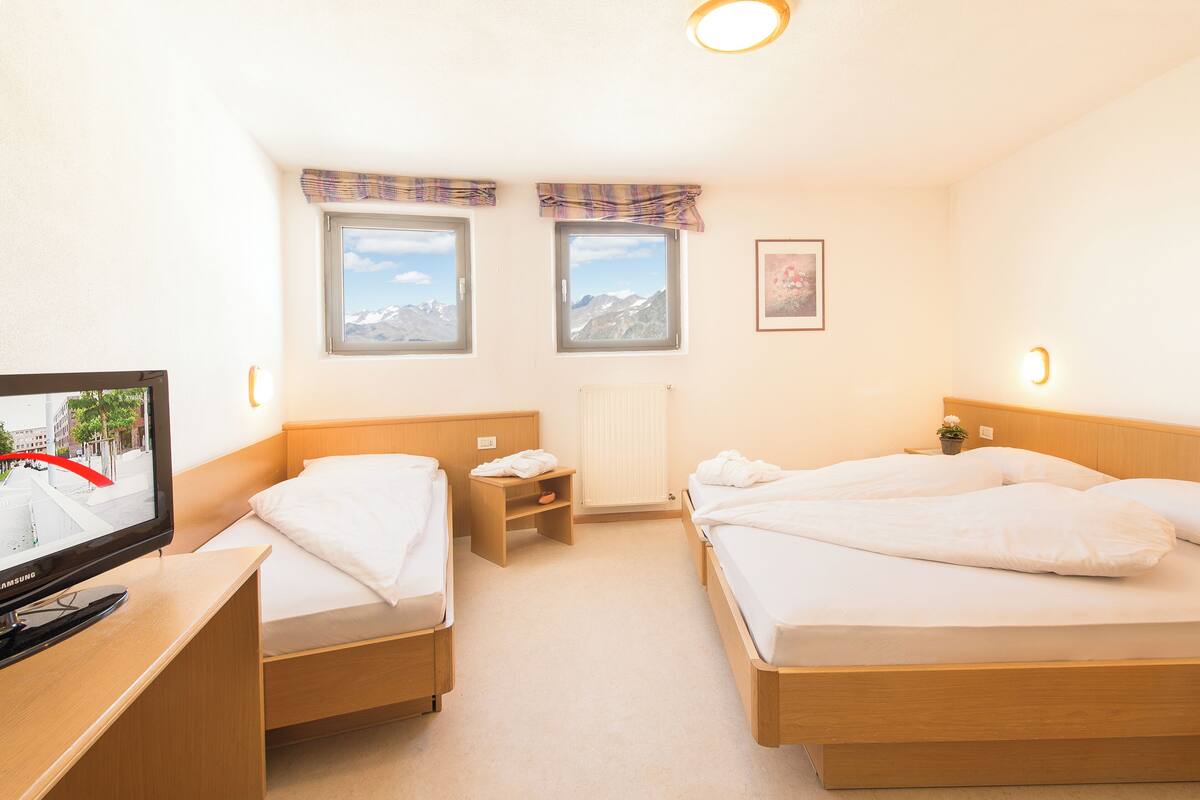 A hotel room with two beds, a TV, a small table, a nightstand, a window with a mountain view, and a radiator.