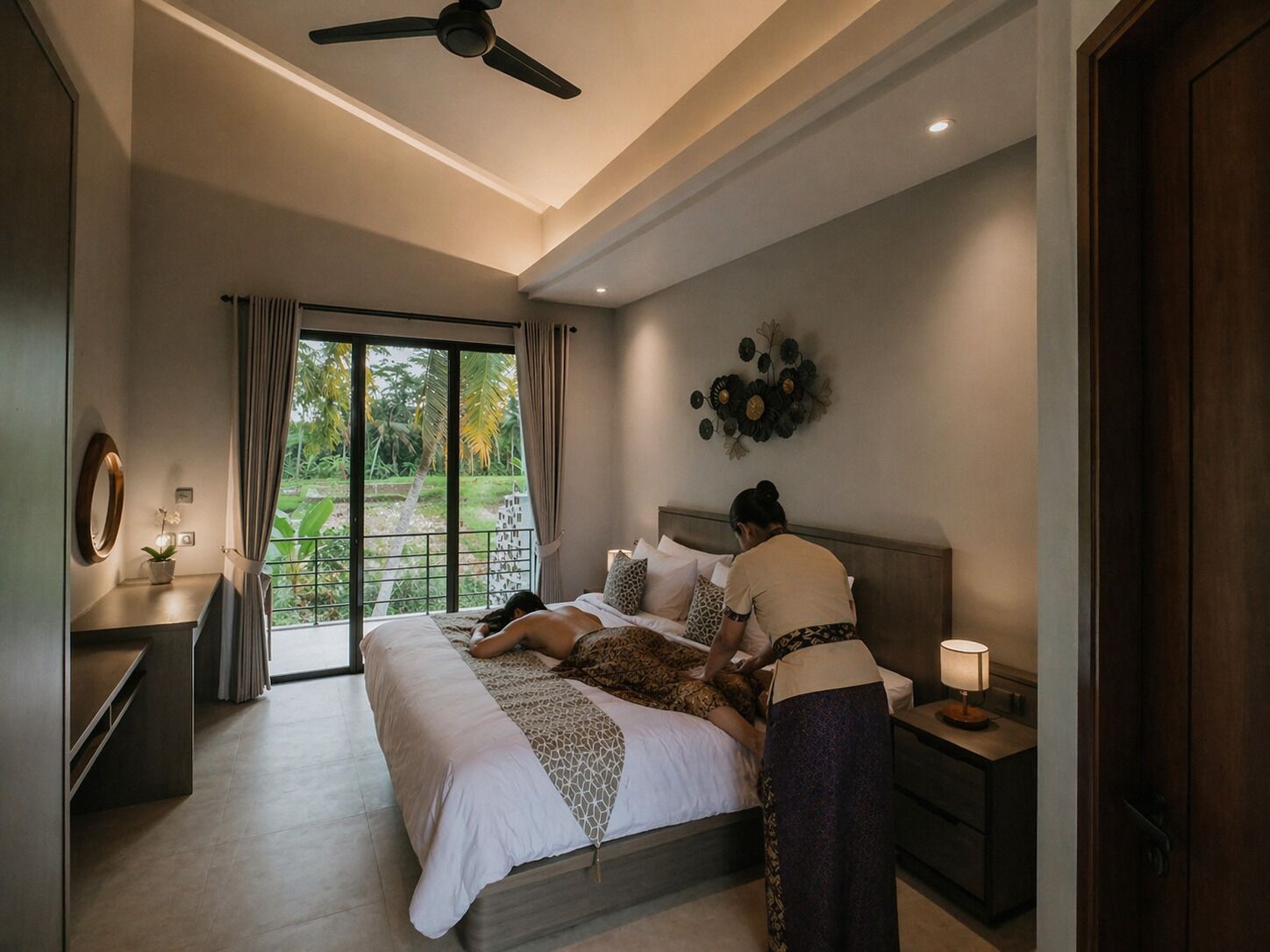 A bedroom with a bed, a nightstand, a lamp, a ceiling fan, and a view of greenery outside.