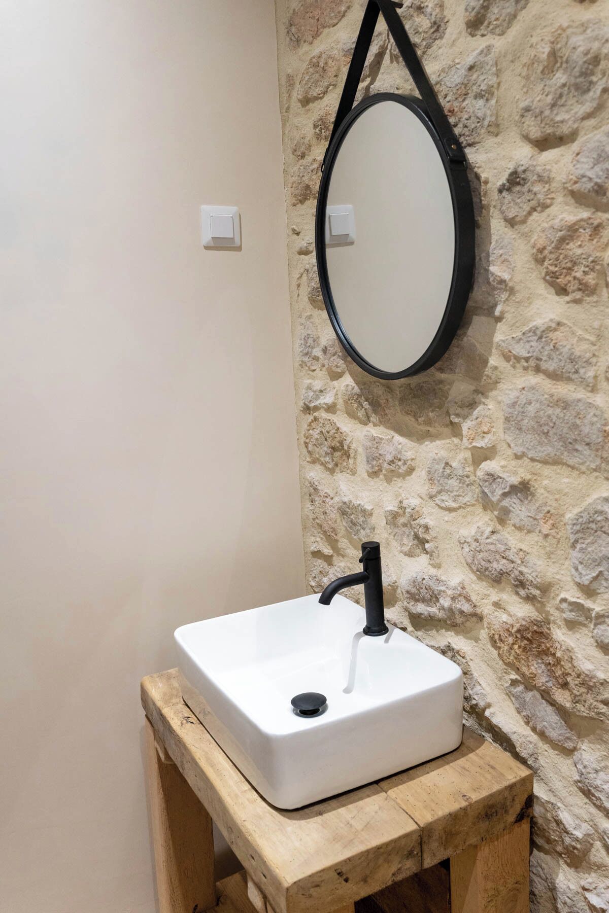 Une salle de bain avec un lavabo blanc posé sur un tabouret en bois, un mur en pierre et un miroir rond.