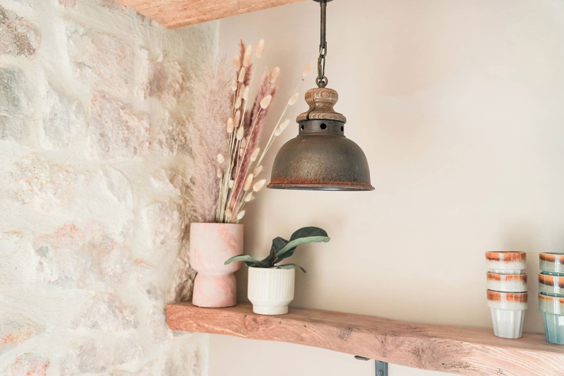 Une étagère rustique ornée d’une lampe métallique suspendue, d’un vase rose contenant des plantes séchées, d’un pot blanc avec une plante verte et d’une pile de récipients en céramique.