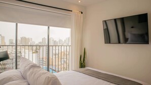 A hotel room with a large bed, a TV mounted on the wall, and a view of a cityscape through a large window. - w Stunning 1BR in Miraflores (Lima)