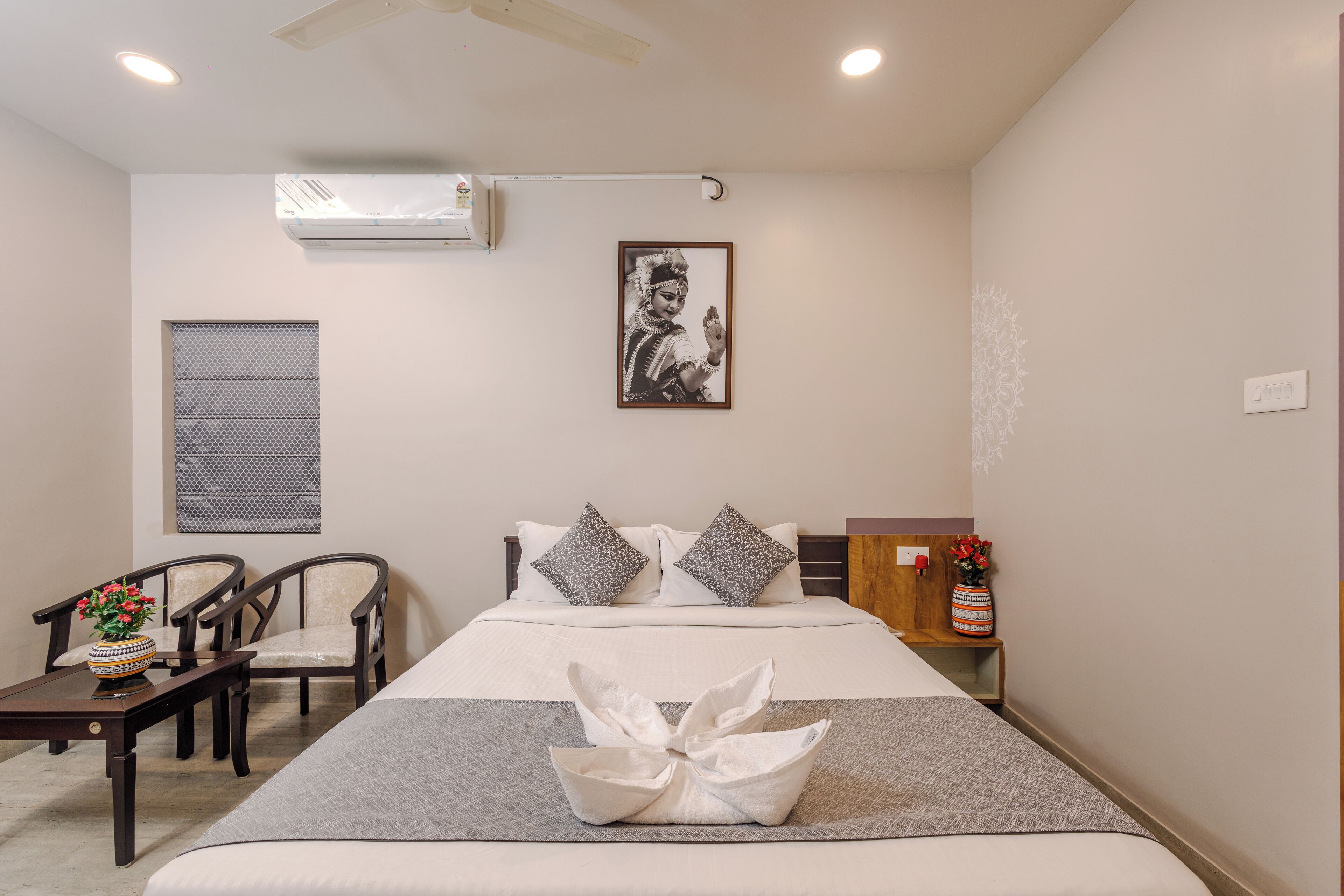 Habitación de hotel con cama, dos sillas, una mesita, un cojín decorativo, aire acondicionado de pared y un cuadro enmarcado.