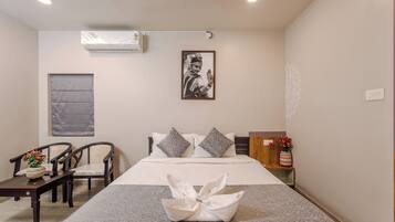 Habitación de hotel con cama, dos sillas, una mesita, un cojín decorativo, aire acondicionado de pared y un cuadro enmarcado.