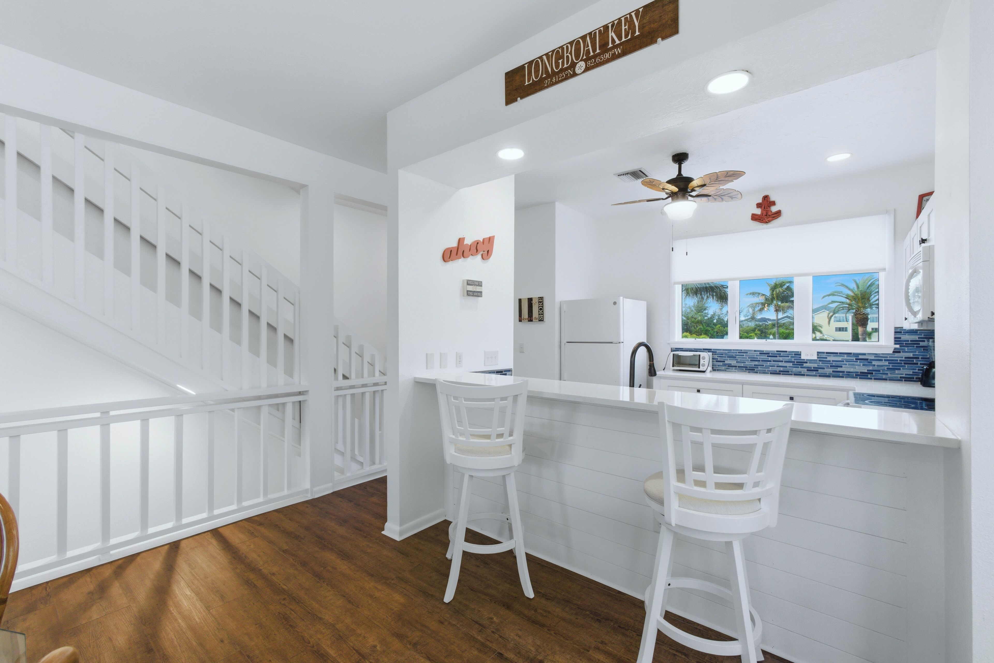 A modern kitchen with white cabinetry, a bar area with white stools, a ceiling fan, and a view of palm trees through large windows.