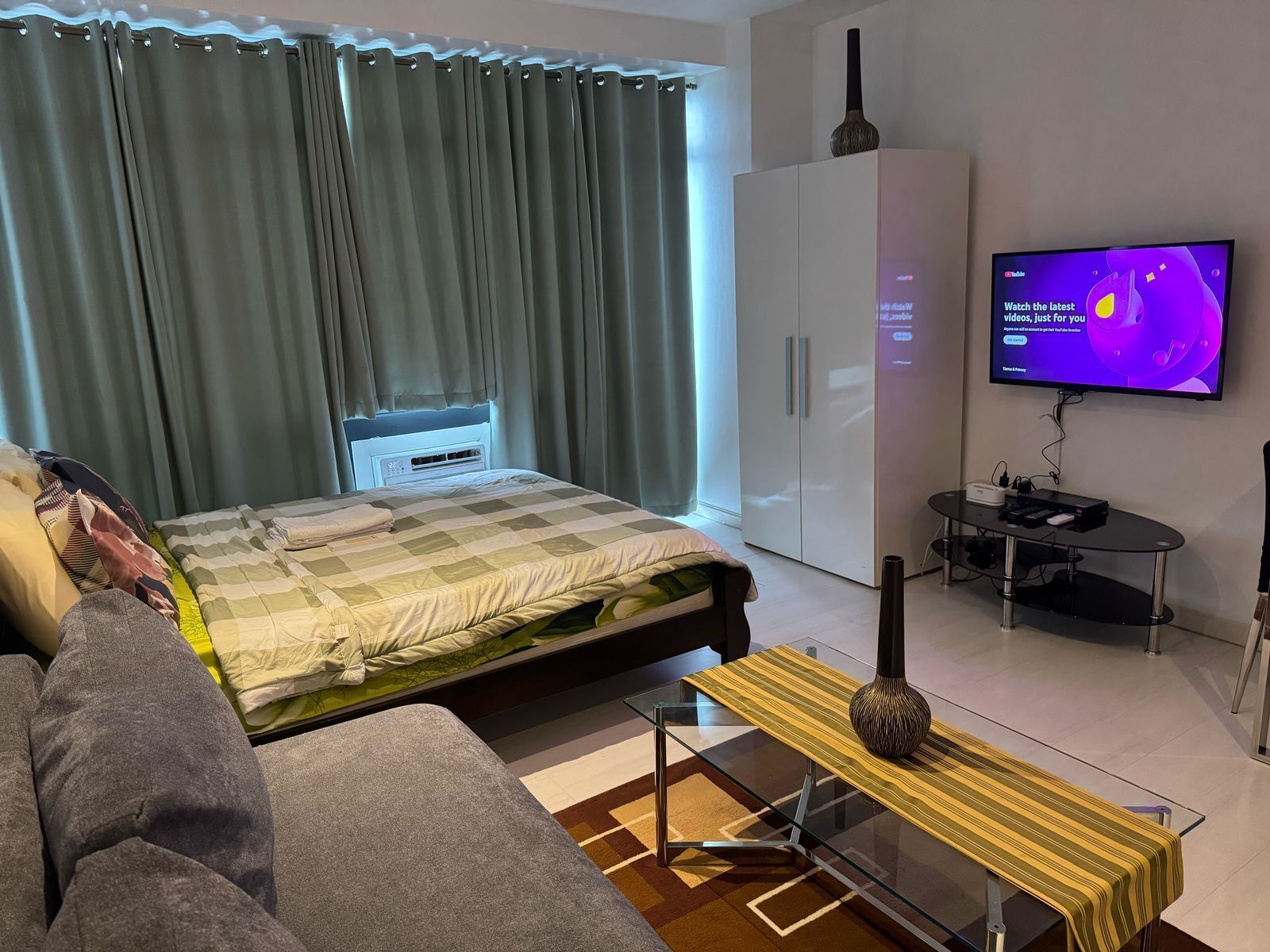 A modern hotel room with a bed, a sofa, a TV, a wardrobe, and a small table with a vase.
