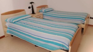 Una habitación pequeña y sencilla con dos camas individuales, cada una con ropa de cama a rayas. Tiene una mesita de noche con una lámpara a un lado y un cajón pequeño al otro. Las paredes son lisas y el suelo está cubierto de baldosas.