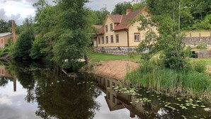A house with a stone foundation and a tiled roof, surrounded by lush greenery and a calm pond with lily pads.