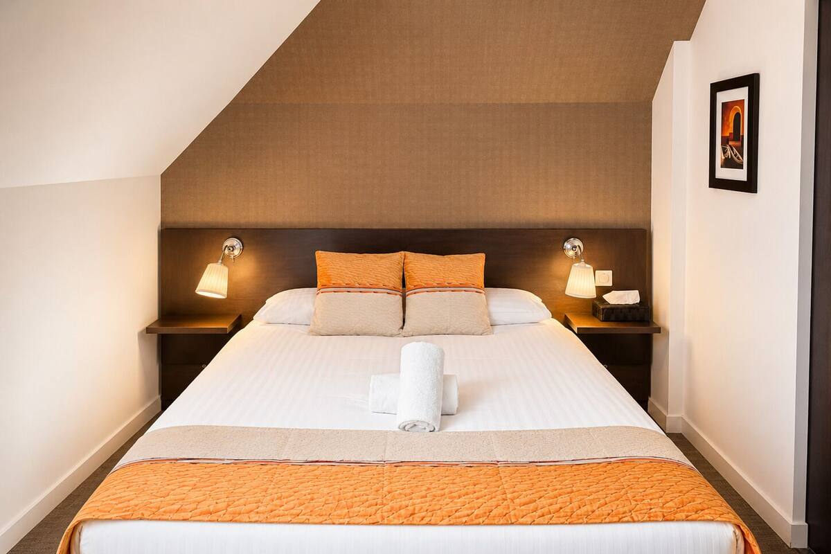 A hotel room with a bed, two bedside tables, a nightstand, a framed picture on the wall, and a towel on the bed.