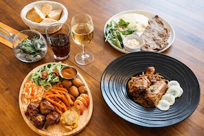 Ein Tisch mit verschiedenen Gerichten, darunter gegrilltes Fleisch, Gemüse und Brot. Außerdem stehen Gläser mit Getränken und eine Schüssel Brot darauf.