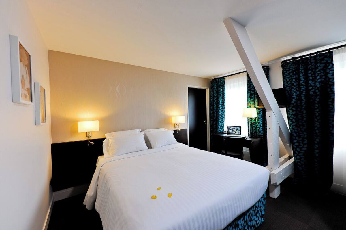 A hotel room with a large bed, two bedside lamps, a desk with a chair, and a window with curtains.
