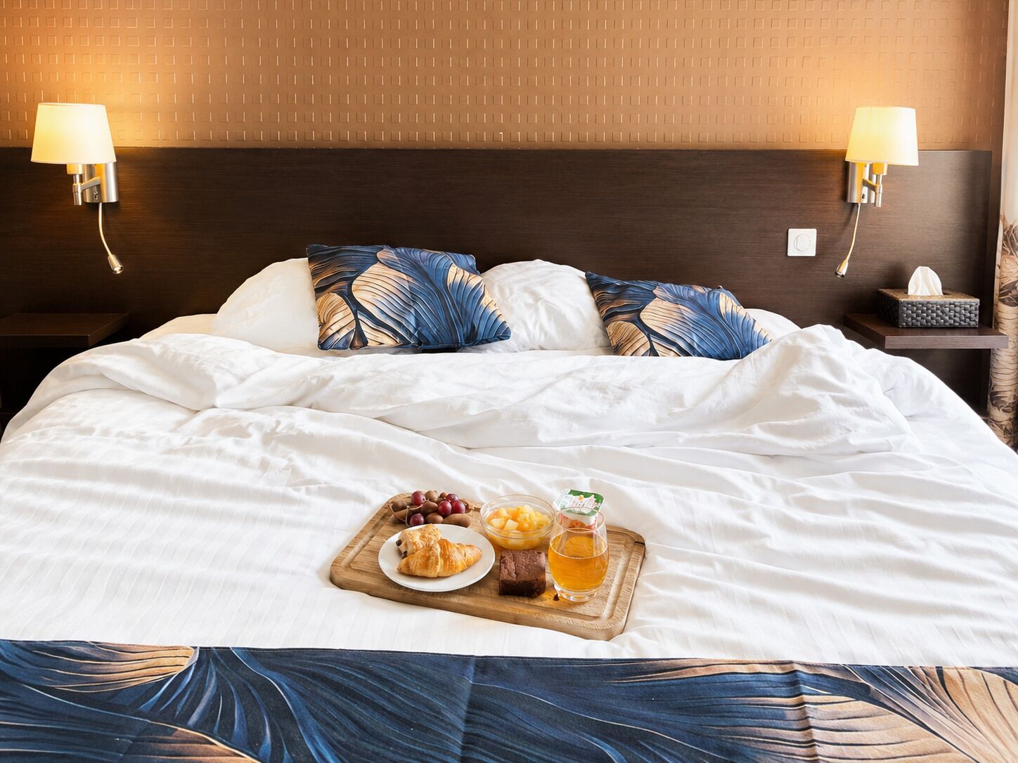 A hotel room with a neatly made bed, two wall-mounted lamps, a bedside table with a tissue box, and a tray with food and drinks.