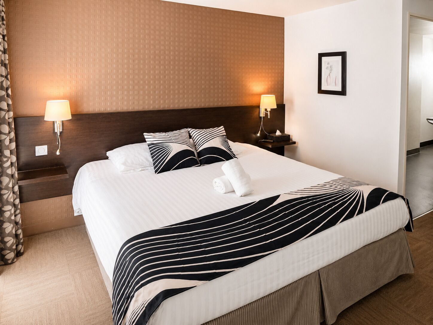 Un lit bien fait, avec une literie à rayures, deux lampes de chevet, un tableau encadré au mur et une tête de lit en bois.
