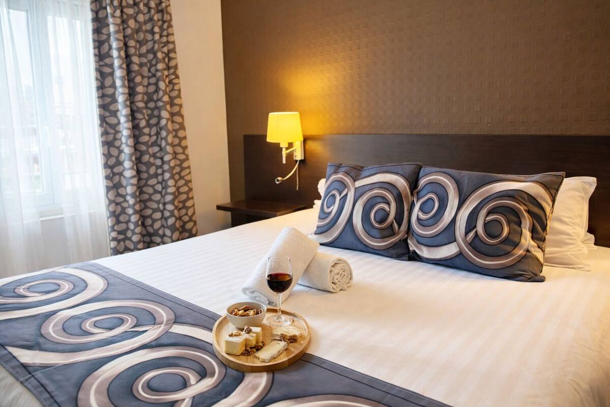A neatly made hotel bed with a patterned bedspread, pillows, a tray with snacks and a glass of wine, and a nightstand with a lamp.