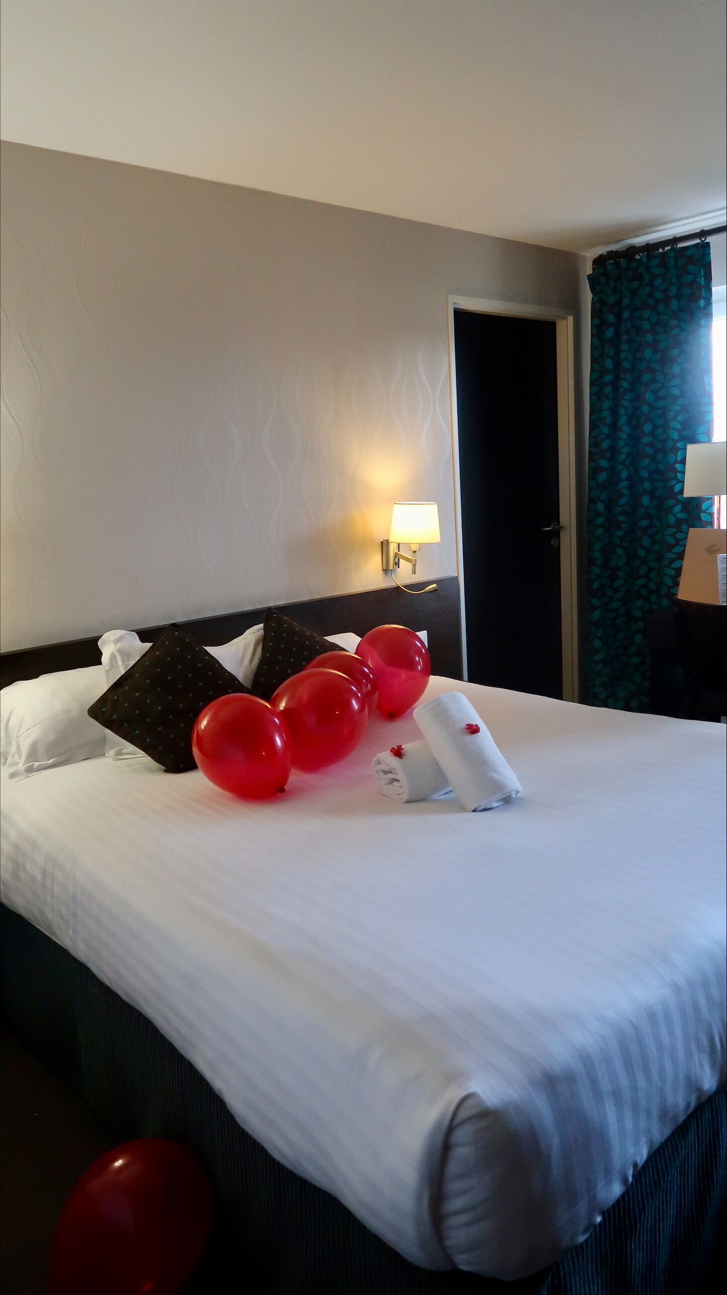 A hotel room with a neatly made bed, red balloons, a folded towel, and a wall-mounted lamp.