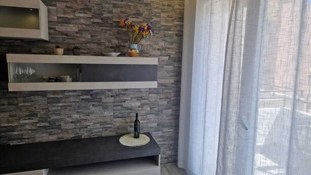 A modern living room with a stone-patterned wall, a floating shelf with decorative items, a dark console table, and a window with sheer curtains. - KLIMT HOUSE. (Marsala)