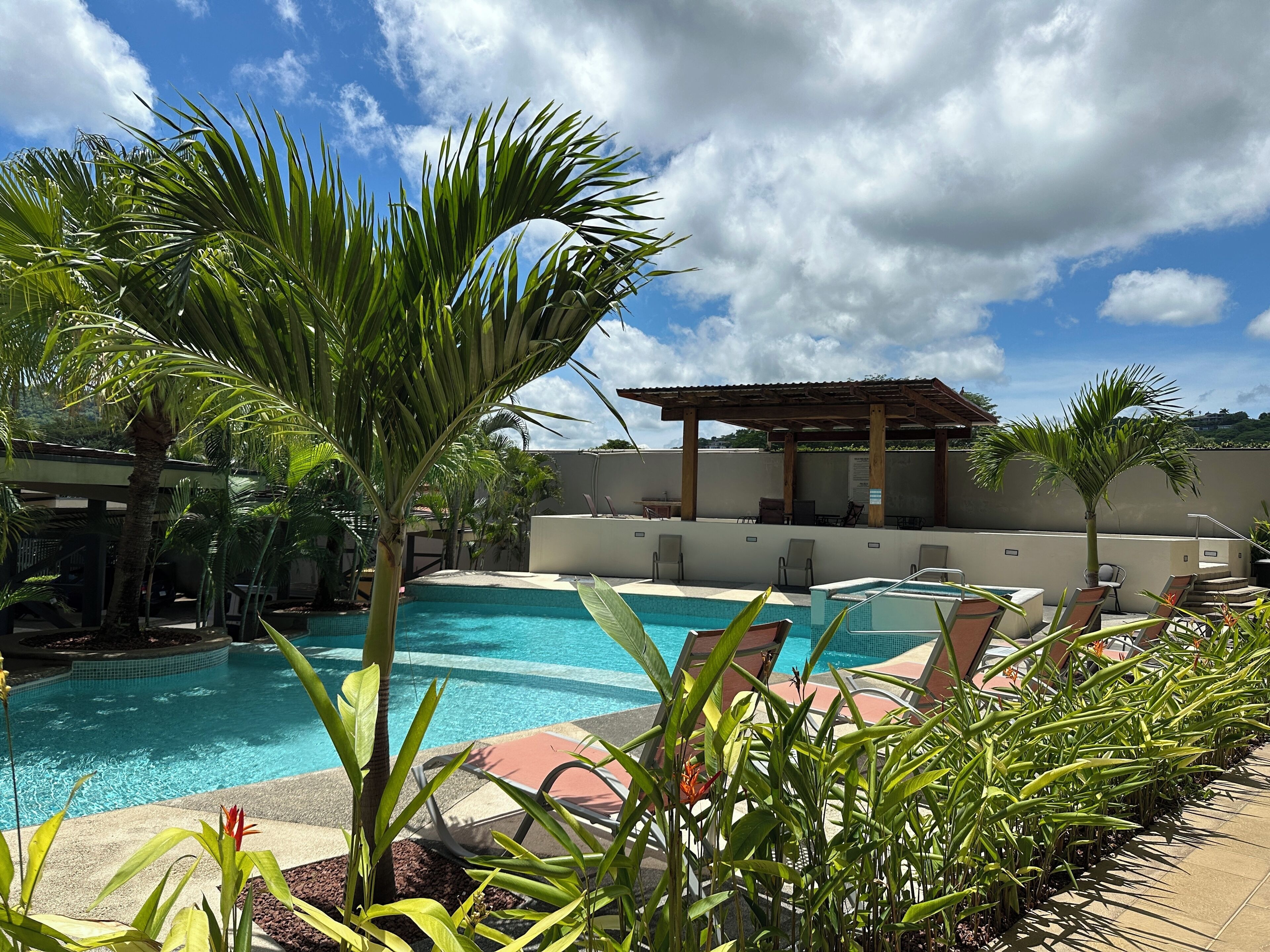 A pool area with lounge chairs and a pergola, surrounded by tropical plants.