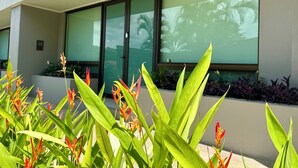 A modern building with large windows reflecting palm trees and a well-maintained garden with green foliage and red flowers. - Exceptional three-bedroom, two-and-a-half-bathroom ground floor condo (Playa Hermosa)