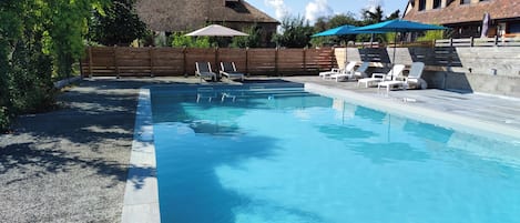 A rectangular swimming pool with clear blue water, surrounded by a wooden fence and a stone wall. There are lounge chairs and umbrellas along the poolside.