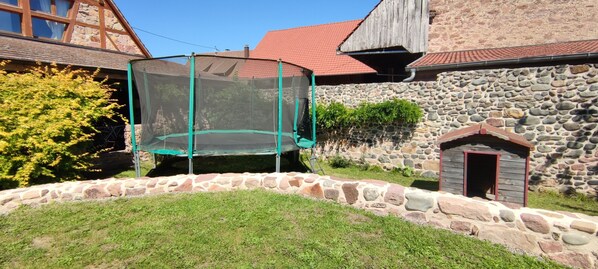 Une maison en pierre avec un toit recouvert de tuiles rouges, un trampoline doté d’un filet de protection et un petit abri en bois.