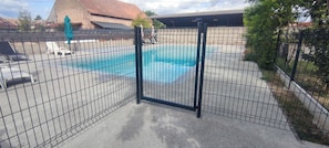 Une piscine entourée d’une clôture, avec des chaises longues et un bâtiment en arrière-plan.