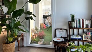 A modern living room with a large sliding glass door leading to a balcony, a bookshelf filled with books, and various potted plants. - Bright apartment with terrace Private parking (Rouen)