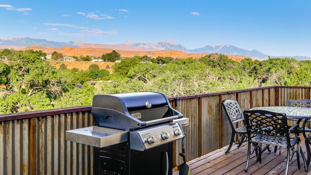 Ein Grill auf einer Holzterrasse mit Tisch und Stühlen mit Blick auf eine gebirgige Landschaft.