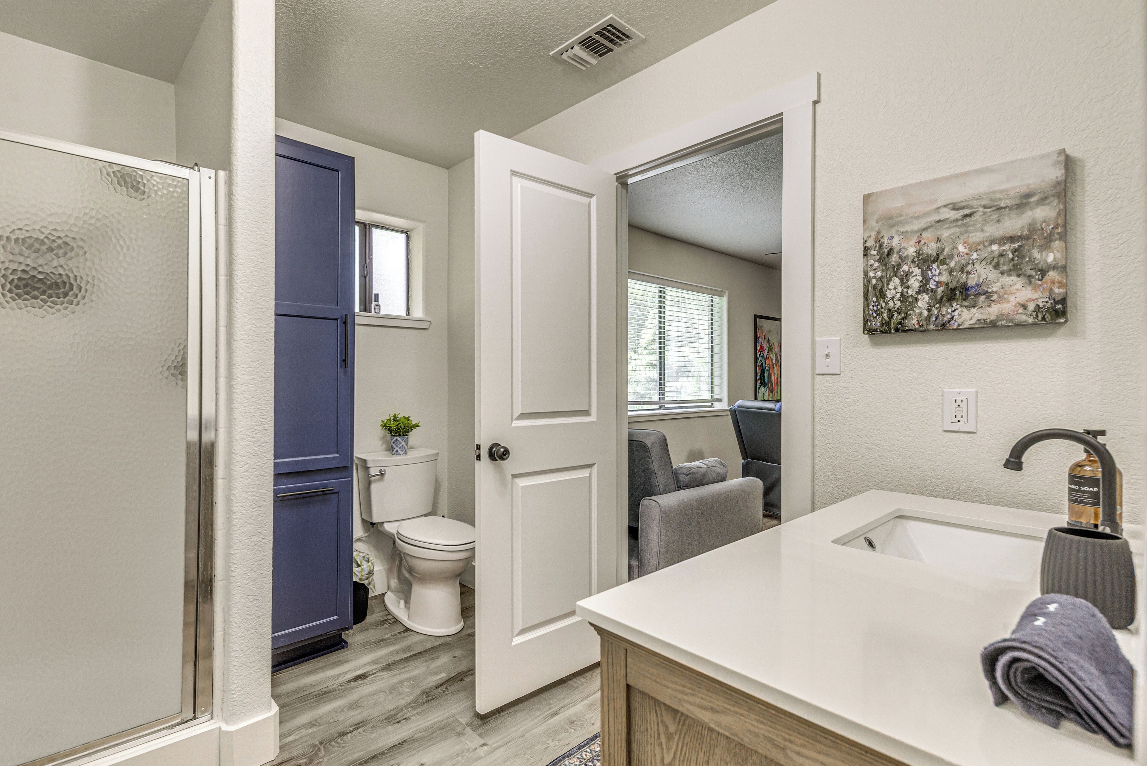 Une salle de bain avec un lavabo blanc, une porte bleue et un tableau accroché au mur.