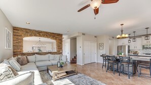 A spacious living room with a stone accent wall, a large sofa, and a coffee table. A kitchen area with bar stools and a chandelier is visible in the background. - Breathtaking Family Retreat in Elite Gold Canyon! (Gold Canyon)