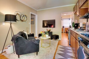 A modern living room with a dark sofa, a floor lamp, and a TV. A kitchen with wooden cabinets and a stove is visible in the background.